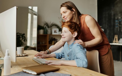 A woman leaning over a seated young boy, helping him on the computer that's on a desk in front of them.  How Neurodiversity-Affirming Assessments Work