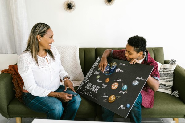 A Black woman and adolescent boy sitting on a couch. The boy is showing the woman a poster about the solar system. What to Expect from a Neurodiversity-Affirming Assessment