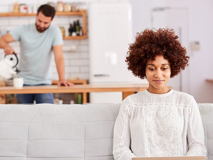 Image of a woman using a laptop while sitting on a sofa, with a man pouring a cup of tea in the background.