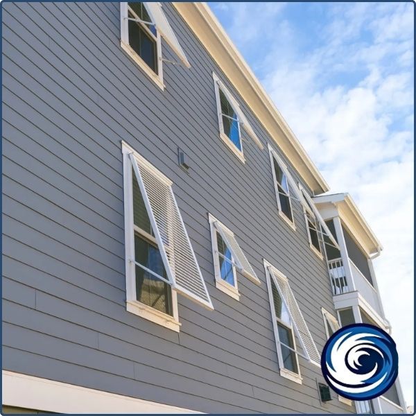 Low-angle view of a grey coastal home with multiple white Bahama hurricane shutters propped open to provide shade and ventilation.