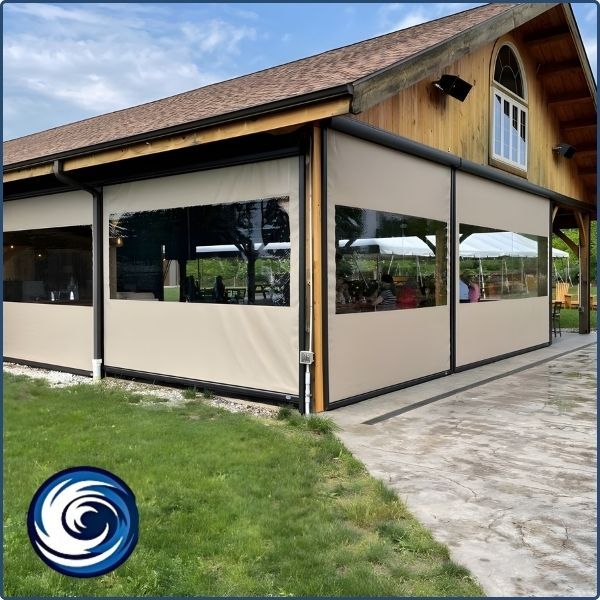 A large outdoor pavilion or restaurant area utilizing heavy-duty clear vinyl weather curtains with tan borders to protect the interior from wind and rain.