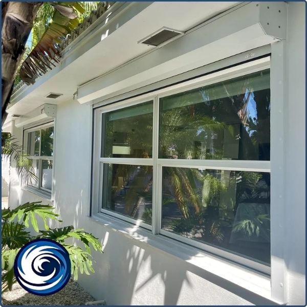 Close-up of white motorized roll-down shutters installed over large glass windows on a modern white home, showing the compact upper housing unit.
