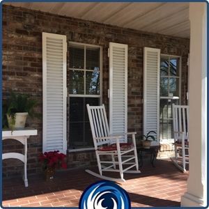 Classic white colonial hurricane shutters installed on a brick house porch, flanking double-hung windows next to a white rocking chair.