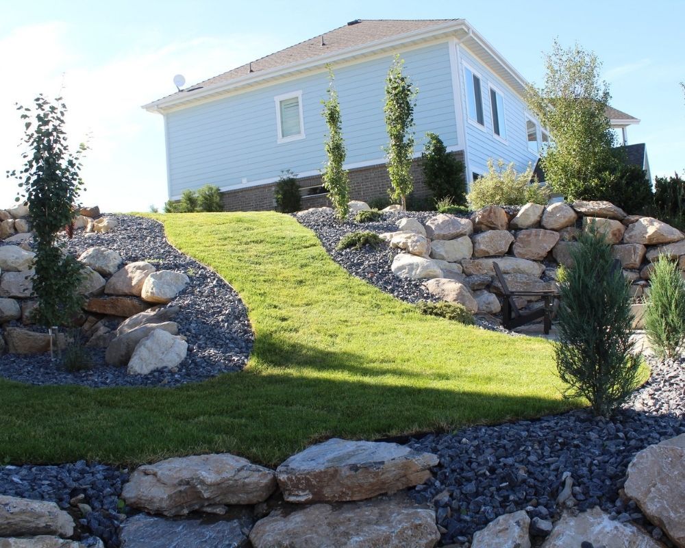 A beautifully sloped backyard landscape featuring a lush grass path, large stone boulders, and decorative privacy trees.