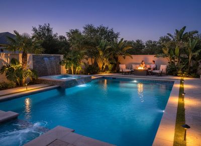 A luxurious backyard features a blue swimming pool, bubbling hot tub, and waterfall. A couple relaxes by a fire pit under lush tropical lighting. Luxurious backyard pool and spa with couple by fire pit at night