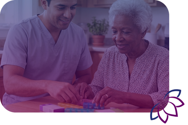 A male caregiver helps an elderly woman organize her daily medications into a colorful pill organizer.