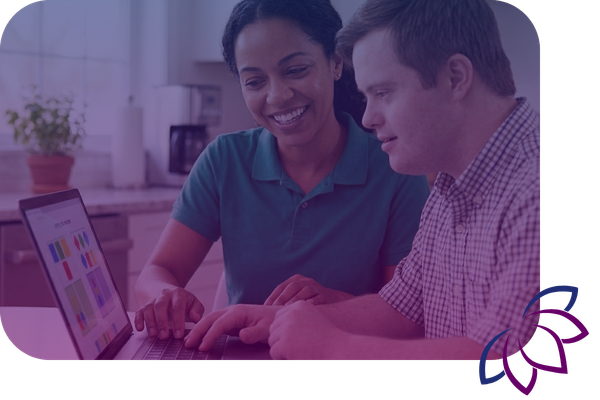 A smiling female caregiver and a young man with Down Syndrome look at a laptop together at a kitchen table.