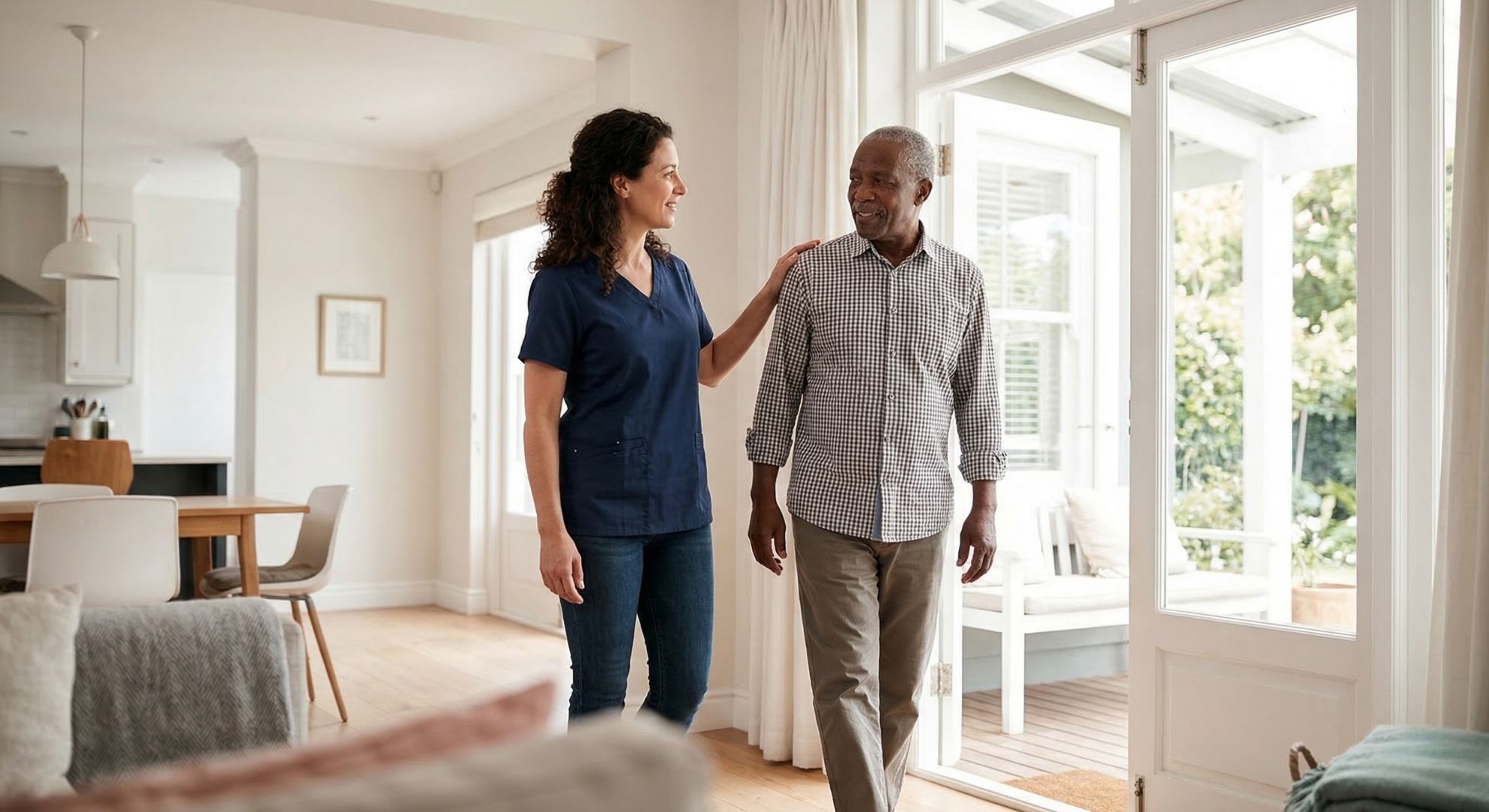 A Lotus Care nurse walks alongside an elderly man in a bright, open-plan home, heading toward a sunlit patio.