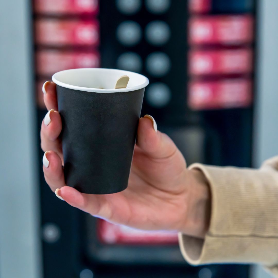 A woman holding up a cup of coffee she got from a vending machine