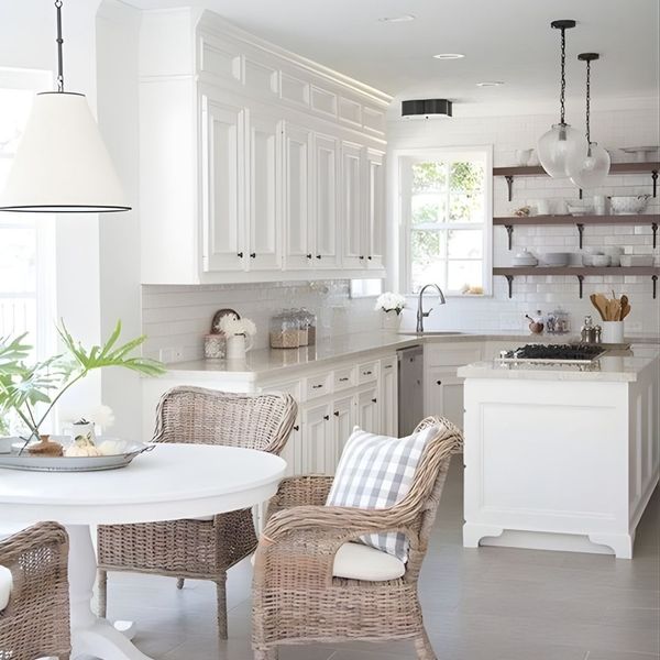 A bright, white coastal-style kitchen featuring woven wicker chairs, white cabinetry, and two different styles of pendant lights hanging over the island and dining table.