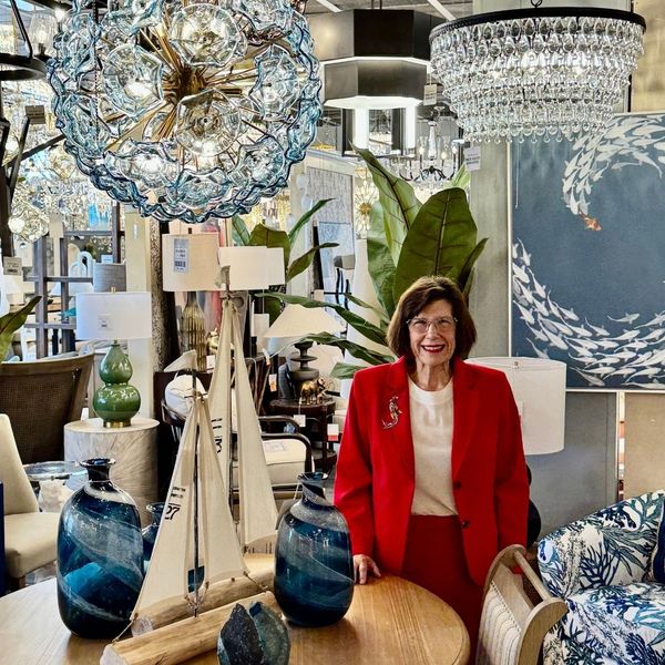 A woman in a red blazer stands in a well-lit showroom surrounded by nautical-themed decor, blue glass vases, and a large, intricate glass orb chandelier.