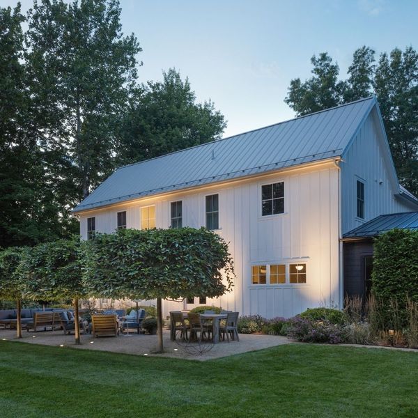 A modern white farmhouse with a large patio area lit by subtle ground lighting and warm glows from the windows under a twilight sky.