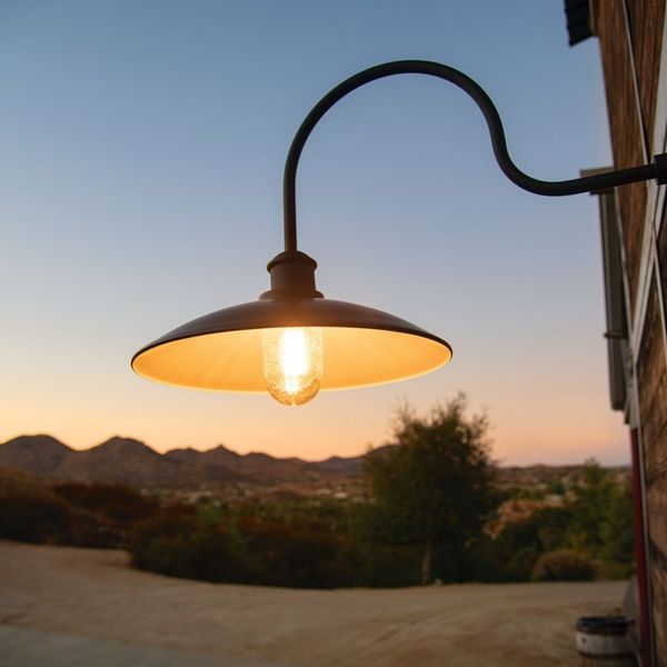 A close-up of a black gooseneck barn light with a warm glowing bulb, set against a sunset backdrop of distant hills.