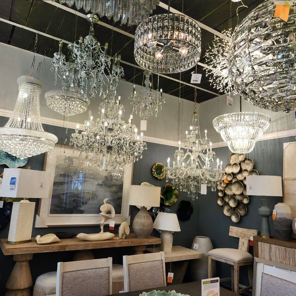 Interior of a lighting showroom featuring a variety of ornate crystal chandeliers and modern hanging lamps above a display of rustic wooden tables and neutral-toned lamps.