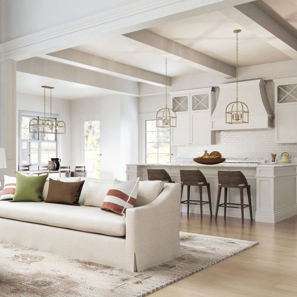 An open-concept living space and kitchen with soft neutral tones, featuring large gold cage-style pendant lights hanging from a beamed ceiling.
