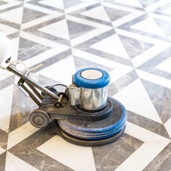 A heavy-duty floor polishing machine with a blue motor and scrubbing pad actively cleaning a black and white geometric patterned stone floor.