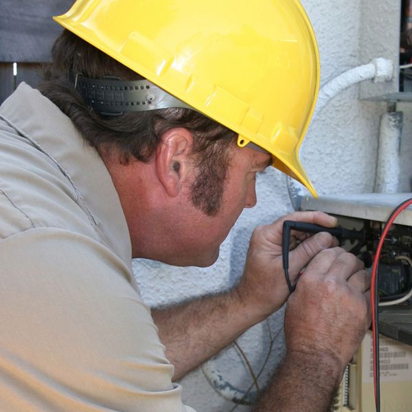 A repair man wearing a yellow helmet testing the voltage on a compressor unit.