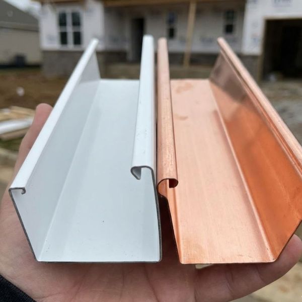 Close-up comparison of white aluminum and copper gutter samples held in hand.