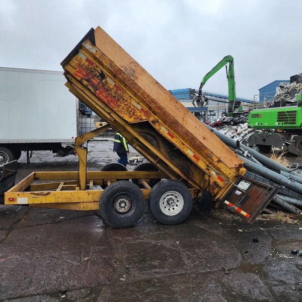 An orange-brown, heavy-duty dump trailer with its bin partially raised, connected to a gooseneck hitch, with other recycling equipment in the background. An orange-brown, heavy-duty dump trailer with its bin partially raised, connected to a gooseneck hitch, with other recycling equipment in the background.