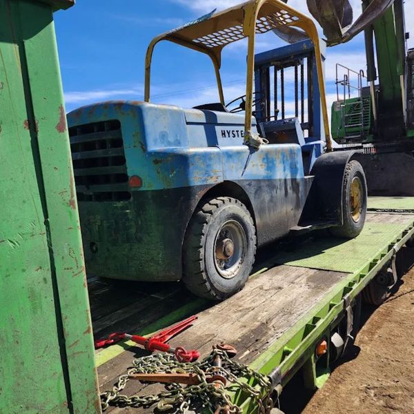 A large, blue-green industrial forklift with thick tires being secured on a flatbed trailer in a sunny, industrial yard. A large, blue-green industrial forklift with thick tires being secured on a flatbed trailer in a sunny, industrial yard.