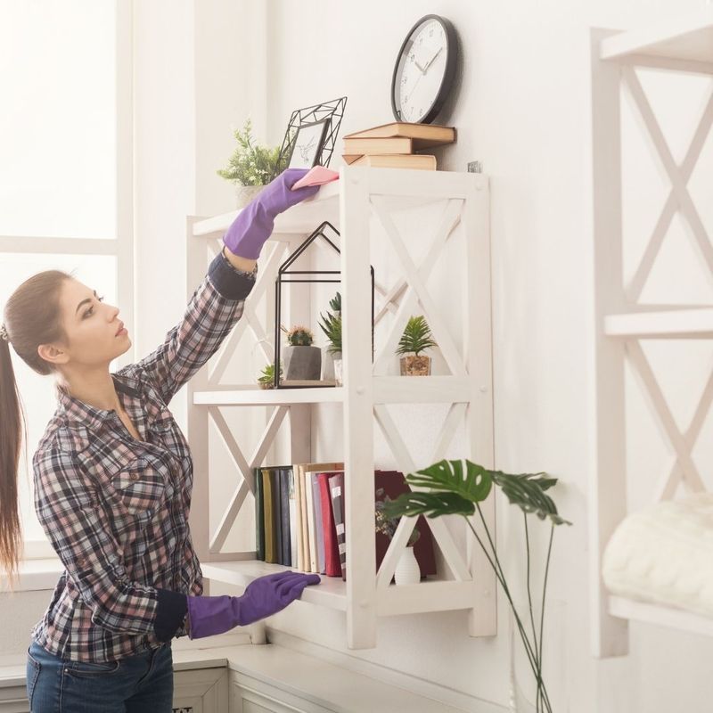 A young cleaning woman dusts the top shelf of a wall fixture