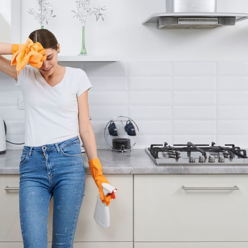 A woman in cleaning cloves holding a cleaning product bottle leans against a counter and wipes forehead