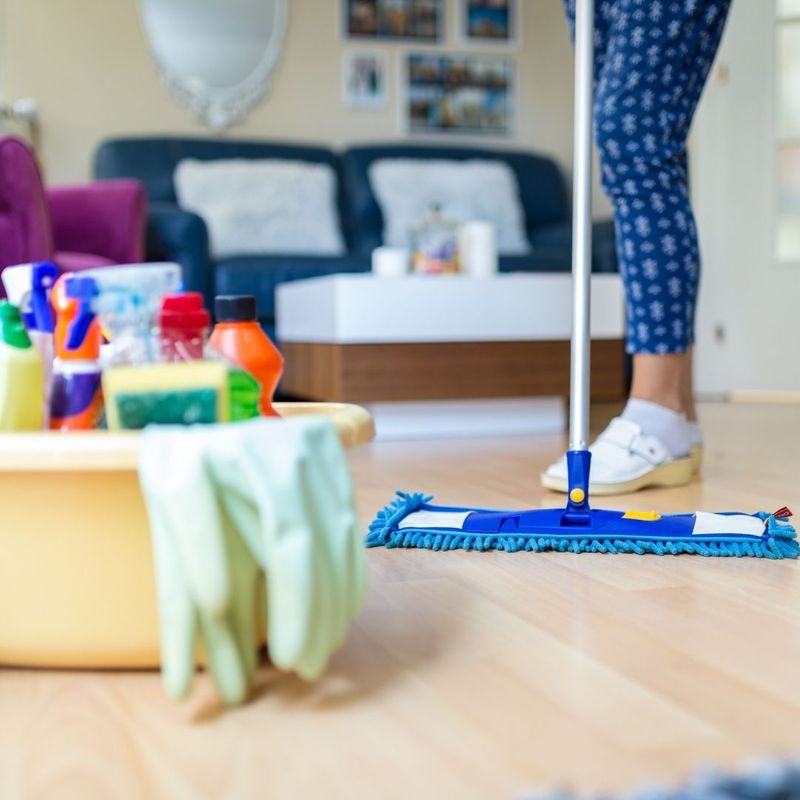 A woman pushes a dust mop across a wood floor next to cleaning supplies