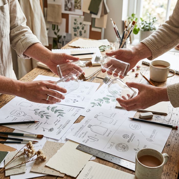 Hands hold several blank, clear glass bottles suitable for sustainable packaging over a workspace with initial label design sketches.