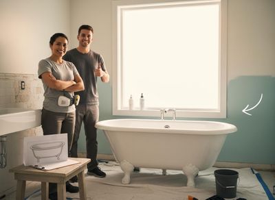 A smiling couple stands in a partially renovated bathroom next to a new white clawfoot tub, with the man giving a thumbs-up. Couple Renovating Bathroom with New Clawfoot Bathtub
