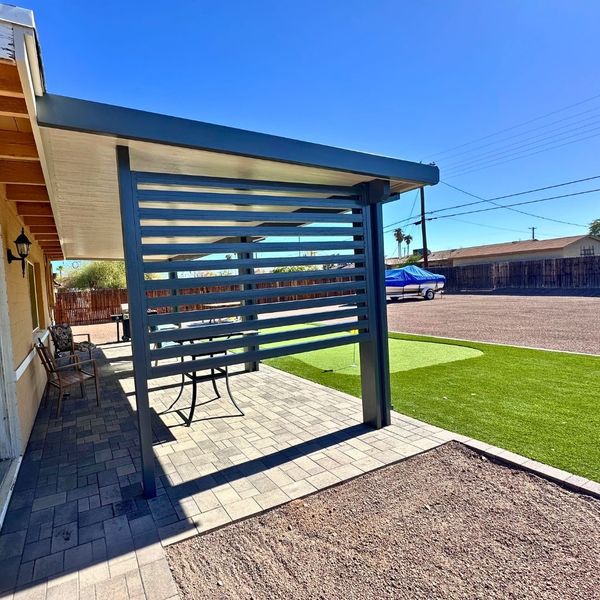 Modern attached solid roof patio cover with a dark grey privacy screen installed over a paver patio and synthetic grass lawn.