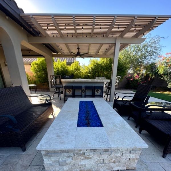 A luxurious outdoor patio features a stone fire pit with blue glass rocks, surrounded by wicker lounge chairs and an outdoor kitchen under a pergola.