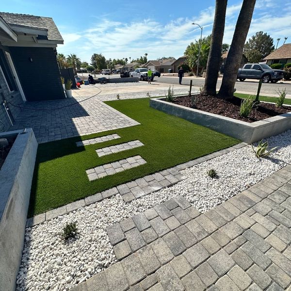 A contemporary front yard features a mix of gray pavers, artificial turf with stepping stones, white gravel, and raised concrete planters.