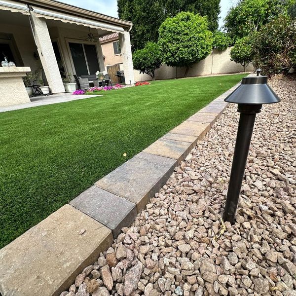 A close-up view of a garden path shows a landscape light next to a stone border separating a gravel bed from a vibrant green artificial turf lawn, with a patio and house in the background.
