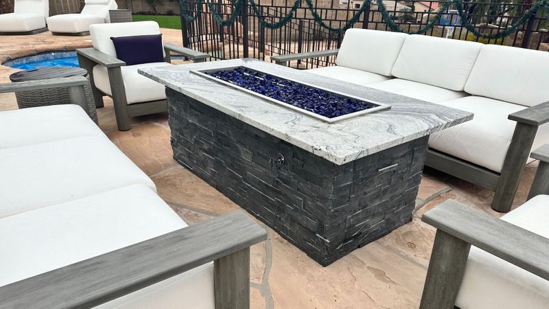 An outdoor seating area showcases a rectangular fire pit with a granite top and a stone base, surrounded by comfortable white cushioned chairs and a sofa on a paver patio.
