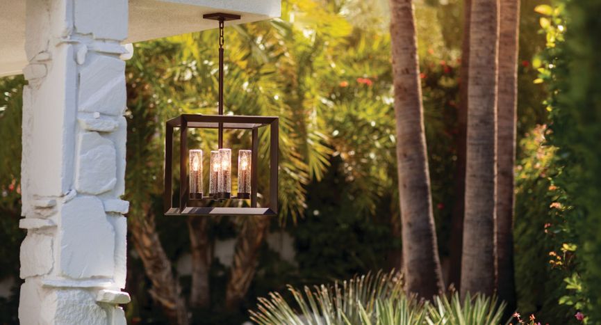 Open-frame bronze hanging lantern with seedy glass cylinder lights suspended from a white patio structure. Open-frame bronze hanging lantern with seedy glass cylinder lights suspended from a white patio structure.