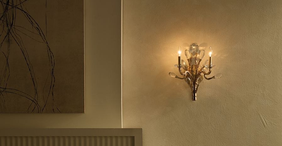 A gold wall sconce with crystal accents and candle-style bulbs mounted on a textured beige wall beside a framed abstract painting.