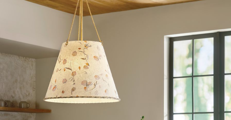A close-up of a tapered pendant light featuring a textured, light-colored shade with a pressed botanical leaf and stem design, suspended from the ceiling by thin brass chains.