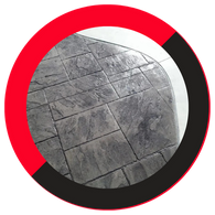 An overhead view of dark grey stamped concrete pavers with a stone texture, bordered by a smooth, light concrete edge in a curved design, framed by a red and black circular border. An overhead view of dark grey stamped concrete pavers with a stone texture, bordered by a smooth, light concrete edge in a curved design, framed by a red and black circular border.