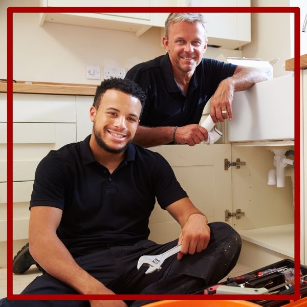 Two friendly Plumbing Plus technicians, a lead plumber and an apprentice, smiling while working together on a kitchen sink installation. Two friendly Plumbing Plus technicians, a lead plumber and an apprentice, smiling while working together on a kitchen sink installation.