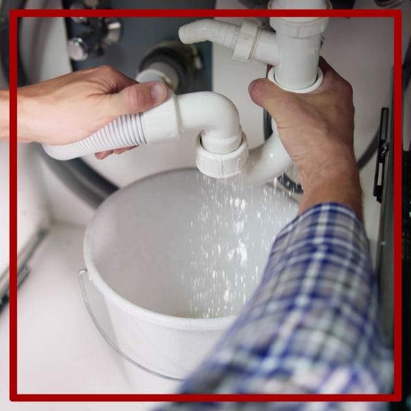 A plumber's hands take control of a major leak from a disconnected white PVC pipe under a sink, with water pouring into a bucket. A plumber's hands take control of a major leak from a disconnected white PVC pipe under a sink, with water pouring into a bucket.