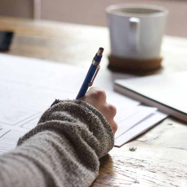 A person wearing a cozy sweater takes notes on paper with a pen, with a mug nearby, suggesting journaling or planning