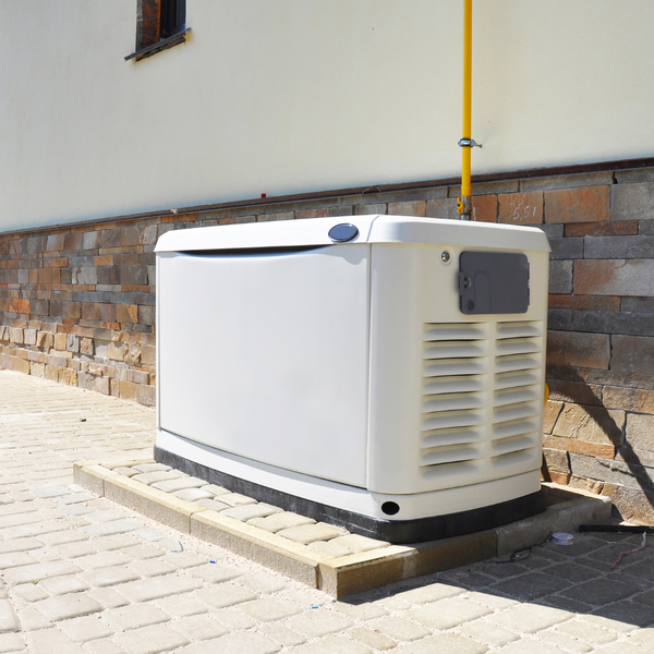 A white, outdoor standby home generator resting on a gravel base next to the corner of a house with a stone and stucco exterior, bordered by green lawn, bushes, and a wooden fence. A white, outdoor standby home generator resting on a gravel base next to the corner of a house with a stone and stucco exterior, bordered by green lawn, bushes, and a wooden fence.