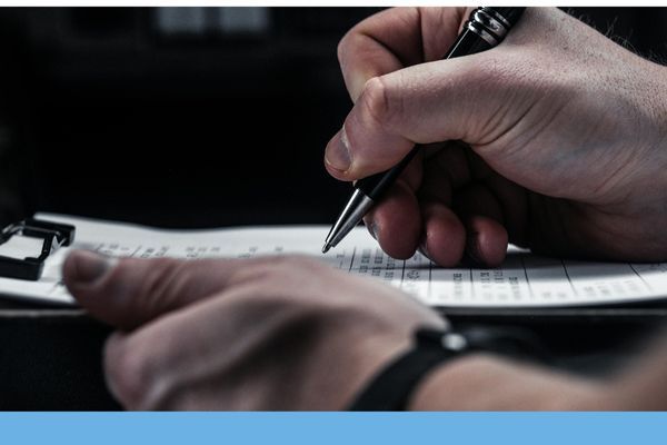 A close-up shot of a person's hands writing with a pen on a checklist or pilot logbook on a clipboard.