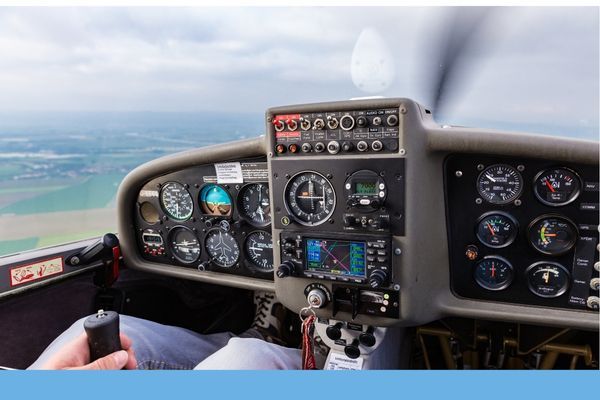 Pilot's view of the cockpit dashboard in a light aircraft, showing analog instruments and a modern GPS display while flying over a green landscape.