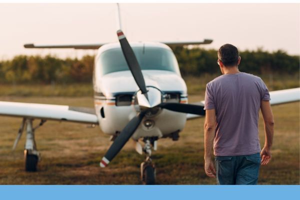 A pilot walking toward a single-engine prop airplane parked on a grassy airfield during golden hour.