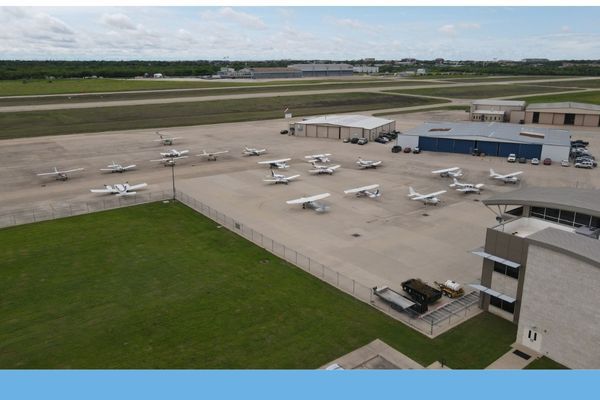 Aerial view of an airport ramp, showing a fleet of single-engine and multi-engine aircraft parked near hangars and a modern terminal building.