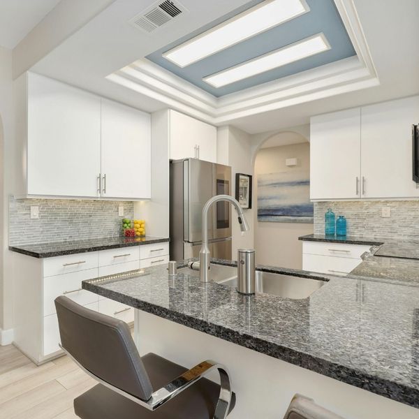 A kitchen with white cabinets and a gray mosaic tile backsplash, featuring dark, speckled granite countertops and a stainless steel sink and faucet.