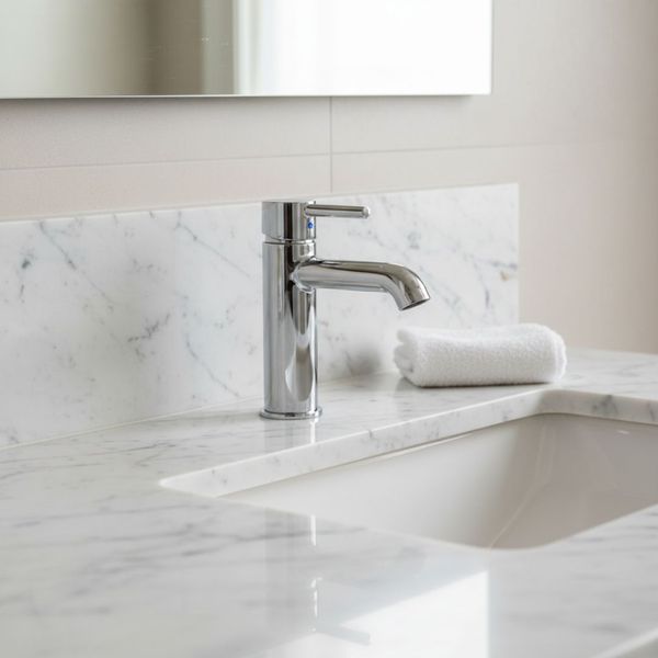 A bright bathroom vanity area featuring a white marble countertop and backsplash with a chrome single-lever faucet and a white undermount sink. A small, rolled white hand towel rests next to the faucet.