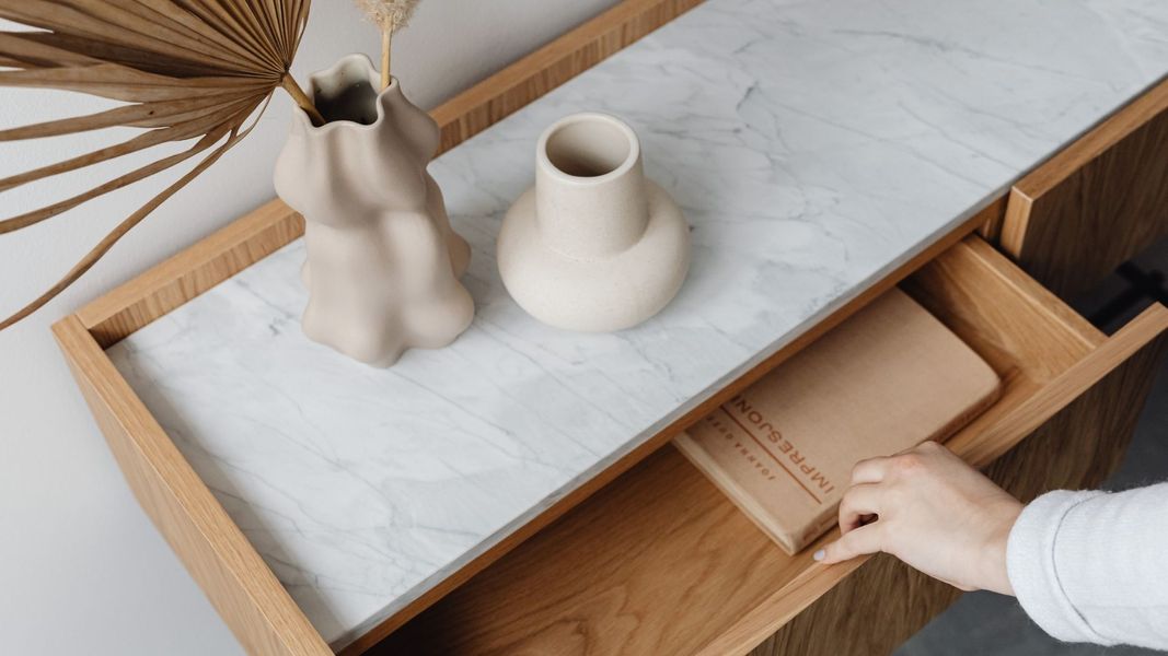 A close-up shot of a modern wooden console or vanity with a section of white marble countertop inset. Two decorative vases and a hand reaching for a notebook inside an open drawer are visible.