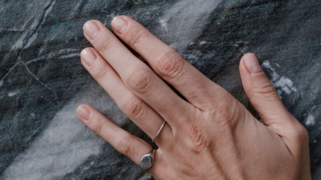 A very close-up detail of a dark, textured natural stone surface, possibly soapstone or marble, showing a person's hand with a silver ring resting on the stone.
